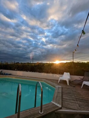 A swimming pool with a wooden deck and lounge chairs, surrounded by a fence and vegetation, under a cloudy sky with the sun setting in the background. - Spacious villa with pool and views of the sea and garrigue (Frontignan)