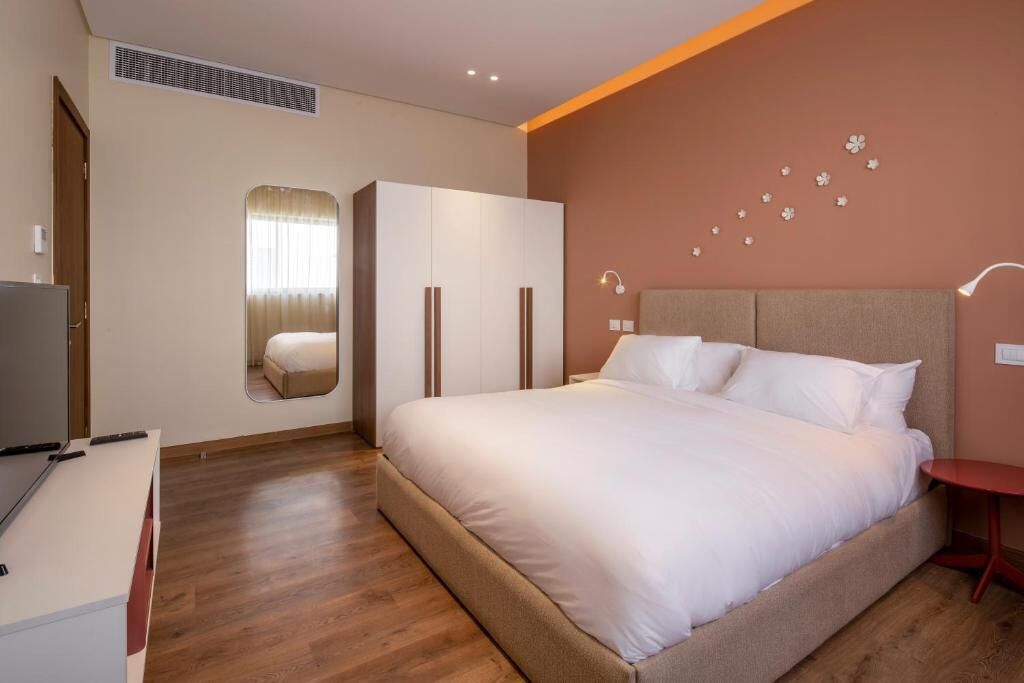 A bedroom with a large bed, a wardrobe, a mirror, a desk, and a red side table.