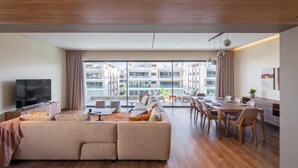 A modern living room with a large sofa, dining table, and chairs. There is a TV on the left and a large window providing a view of the city.