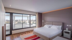 A modern hotel room with a large bed, a view of the city through floor-to-ceiling windows, and a small bedside table.