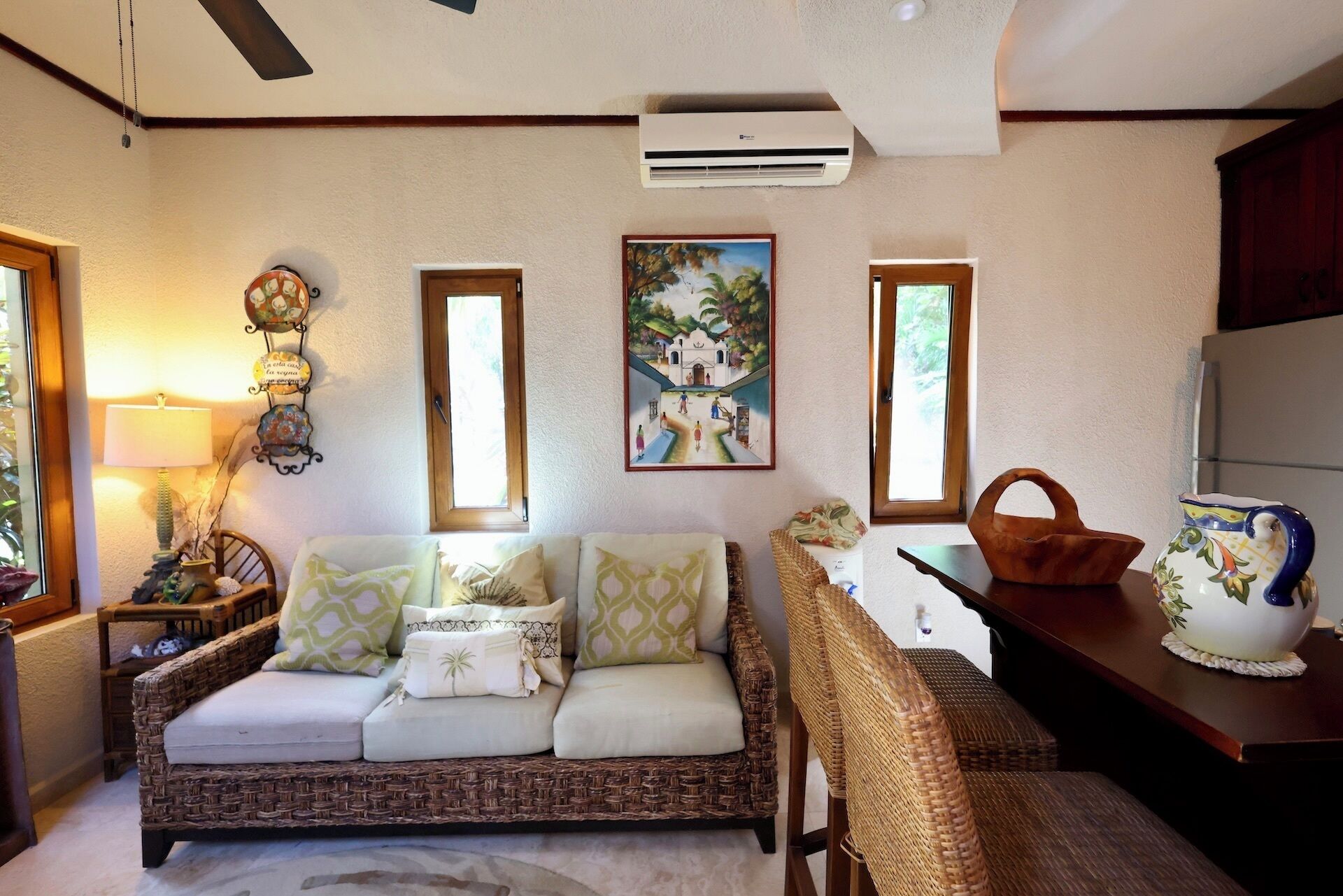 Un salón acogedor con un sofá de mimbre, una mesita con una cesta, una lámpara decorativa y un aire acondicionado instalado en la pared.