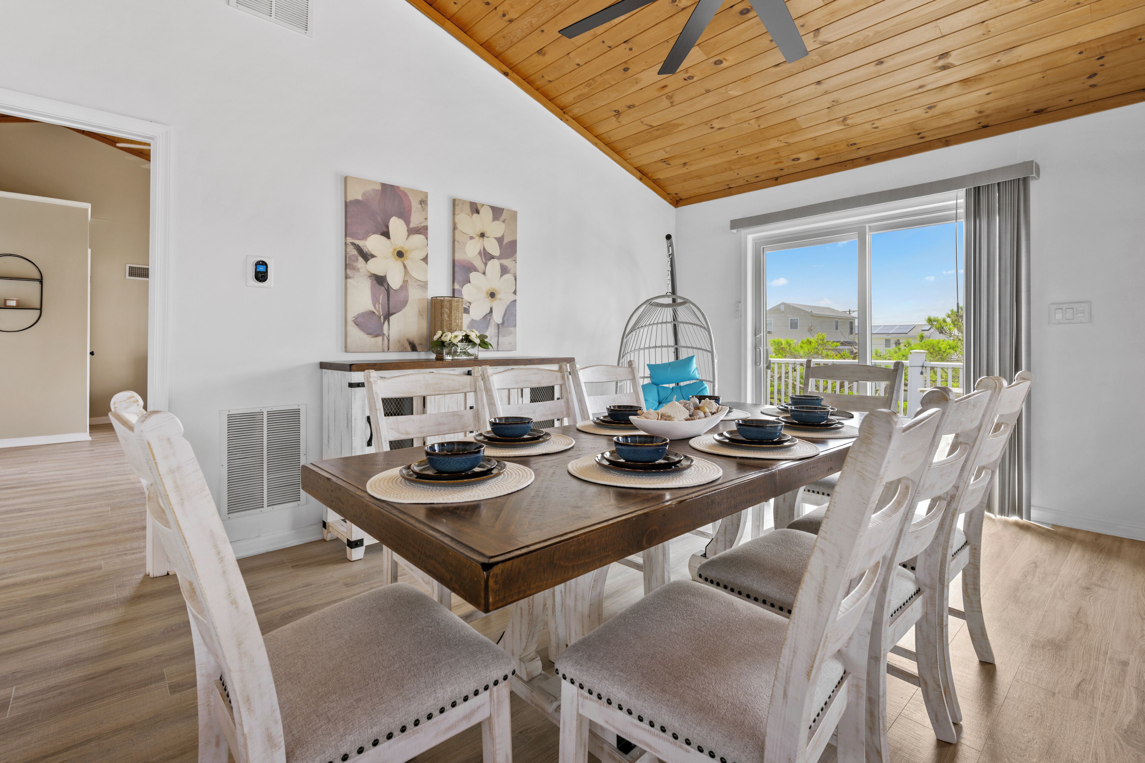 A dining area with a wooden table, white chairs, and a view of the outdoors through large windows.