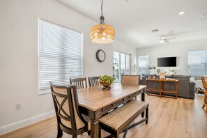 A dining area with a wooden table, chairs, a bench, a hanging light fixture, and a television in the background.