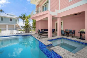 A swimming pool with a tiled edge, a covered patio area with outdoor furniture, and a pink building with a balcony. - The Pink Pearl: Chic beach house with Gulf access & sunset views—ideal Fort Myers Beach escape! (Fort Myers Beach)