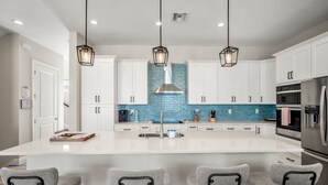 Une cuisine moderne avec des meubles blancs, un îlot central, des tabourets de bar, des luminaires suspendus et des appareils électroménagers en acier inoxydable.