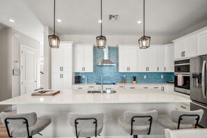 A modern kitchen with white cabinets, a central island, bar stools, pendant lighting, and stainless steel appliances. - The Pink Pearl: Chic beach house with Gulf access & sunset views—ideal Fort Myers Beach escape! (Fort Myers Beach)