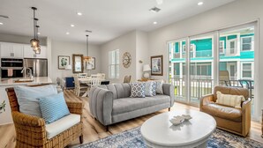 Un salon moderne avec un canapé gris, des fauteuils en osier, une table basse blanche et un balcon donnant sur des bâtiments aux couleurs turquoise.