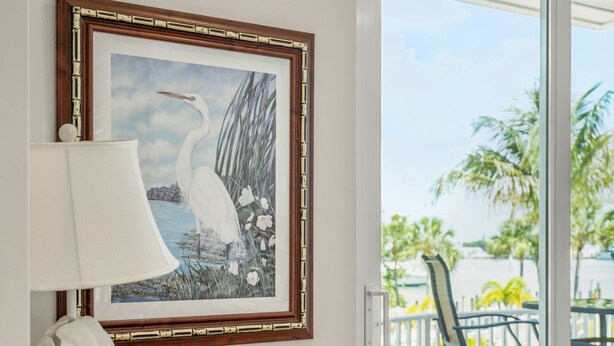 Une pièce avec un fauteuil en osier, un tableau encadré représentant un oiseau et une lampe sur une table.