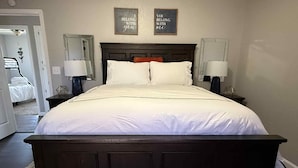 Un dormitorio con una cama grande, dos mesitas de noche y dos lámparas de pared. Hay dos letreros enmarcados en la pared sobre la cama.