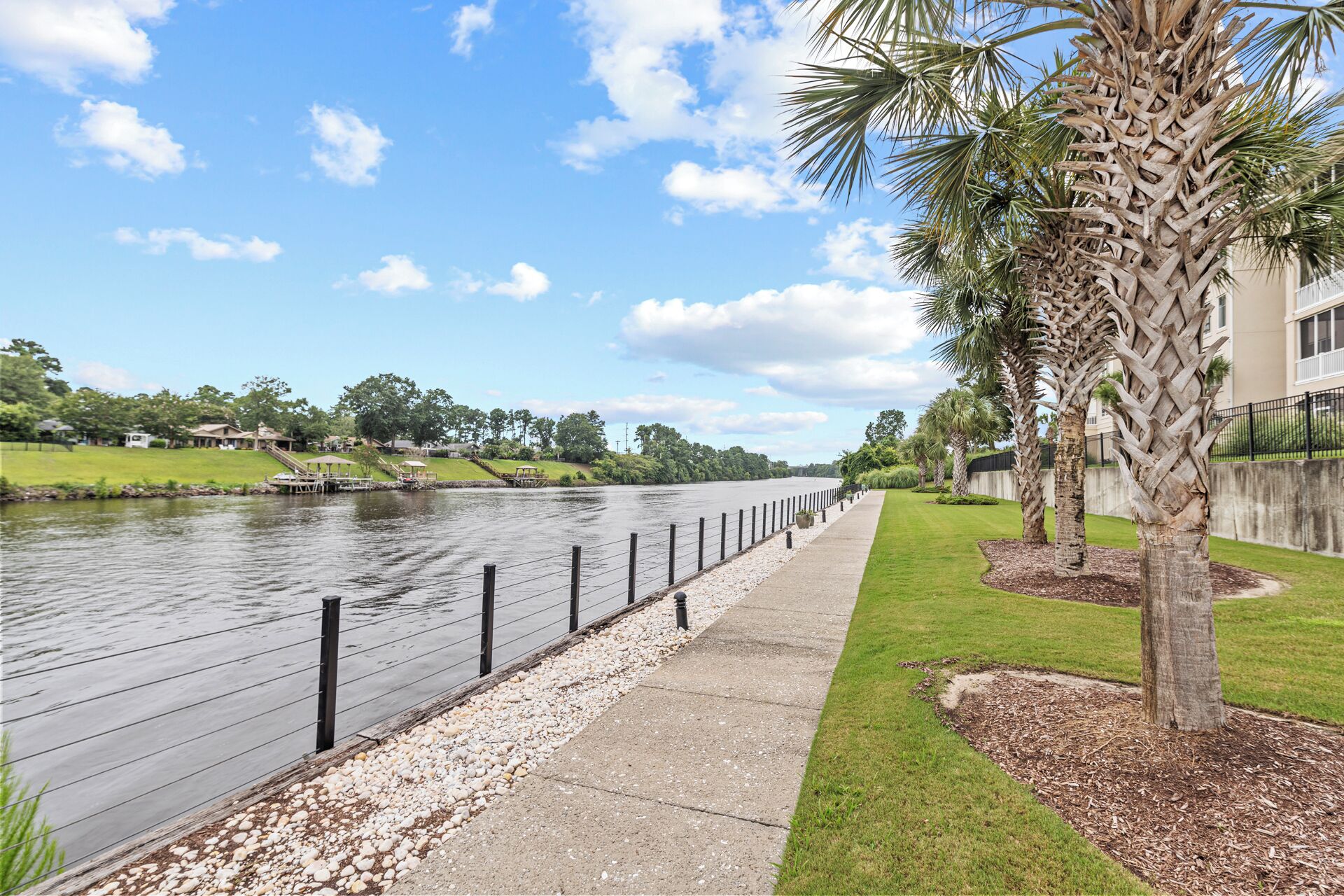A riverside walkway with a fence, pebbles, and palm trees.