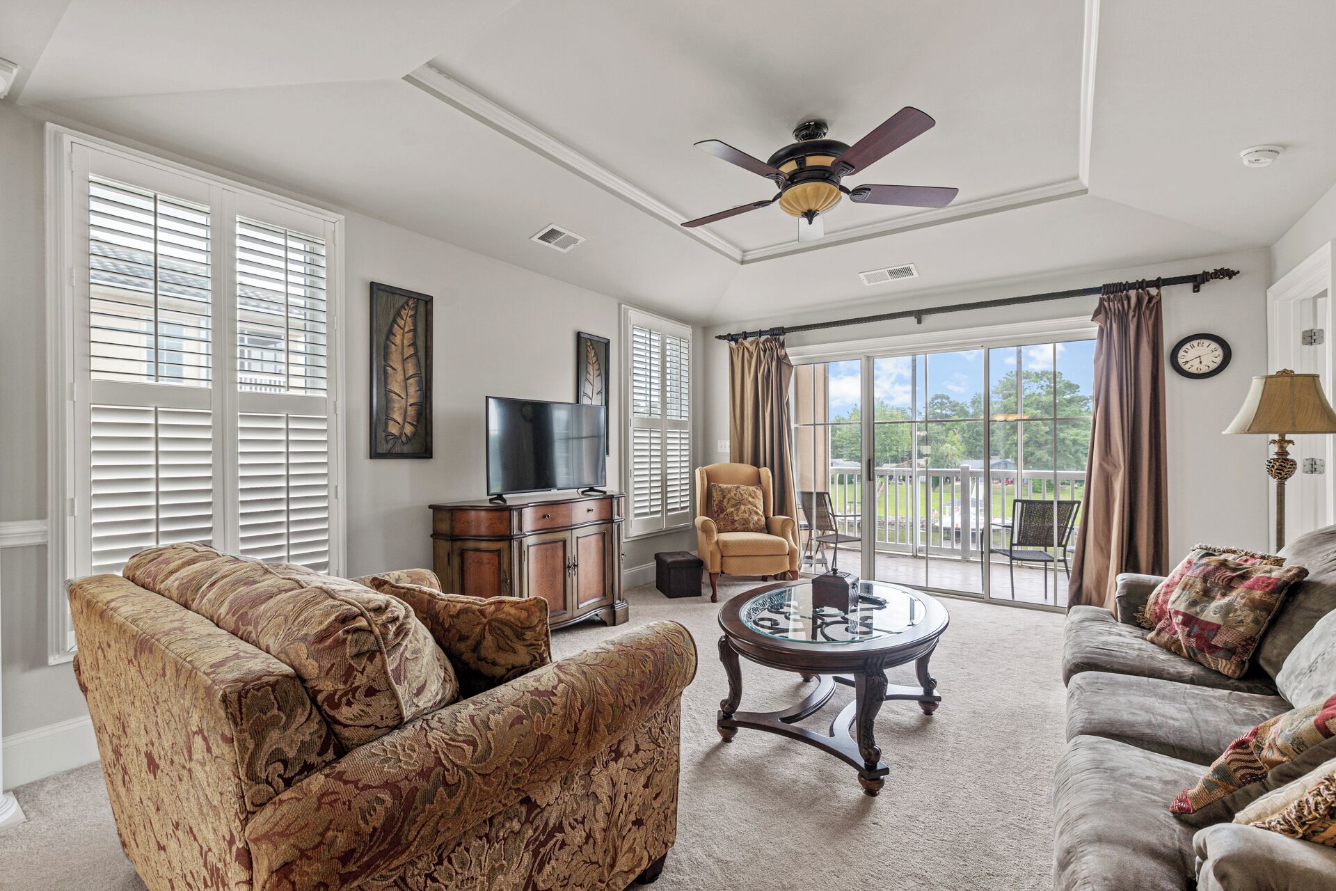 A spacious living room with a ceiling fan, large windows with blinds, a television, and a view of the outdoors.