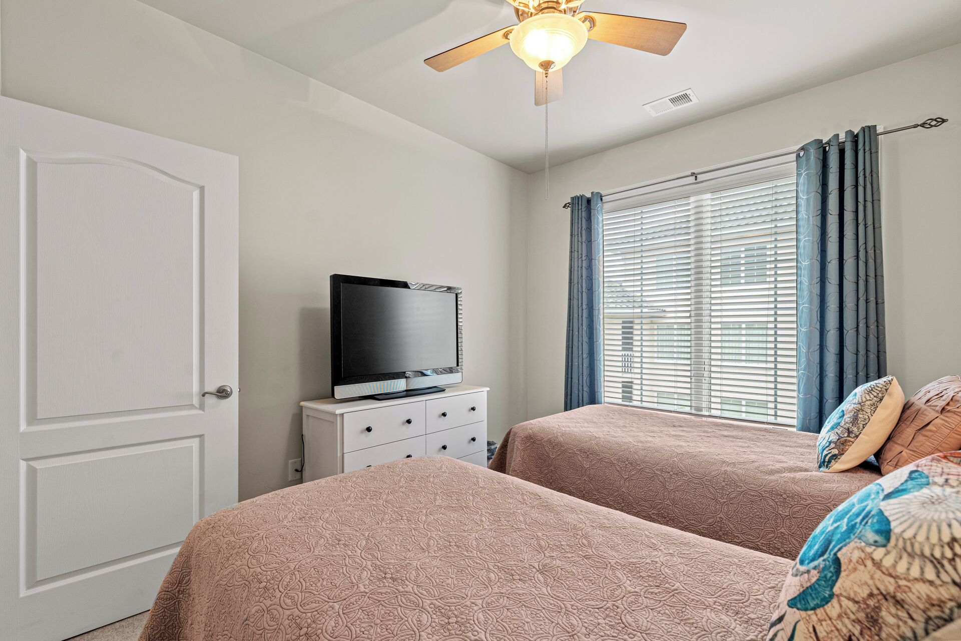 Une chambre d’hôtel avec deux lits, un téléviseur posé sur une commode, un ventilateur de plafond et une fenêtre avec des stores.