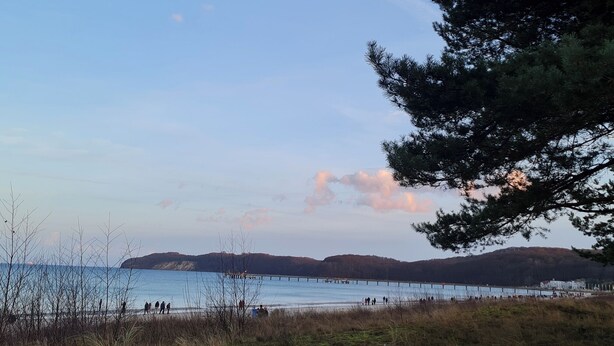 A coastal landscape with a pier extending into calm waters, surrounded by hills and sparse vegetation.