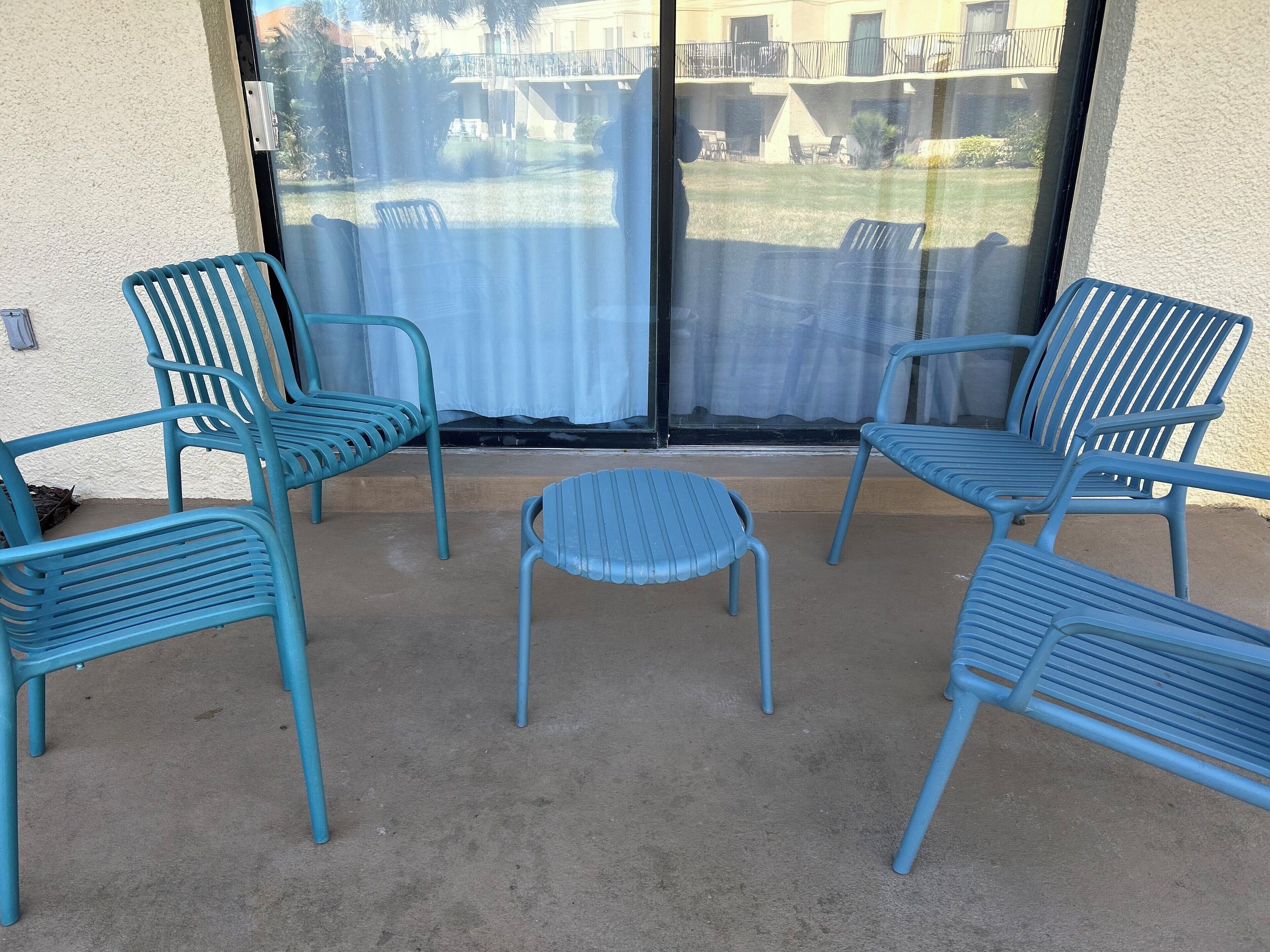 Un patio con cuatro sillas azules y una pequeña mesa, una puerta corrediza de vidrio y vistas a un edificio con mesas al aire libre.