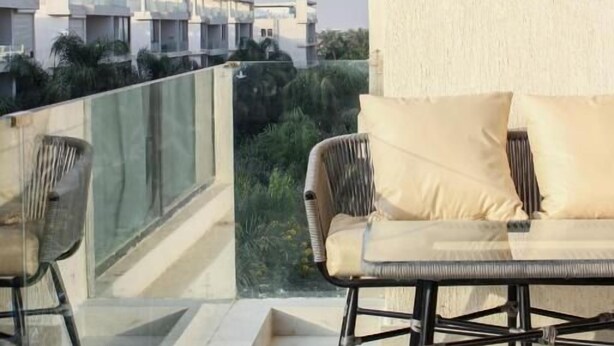 Un balcon avec un canapé et des fauteuils, une rambarde en verre et une vue sur les immeubles voisins.