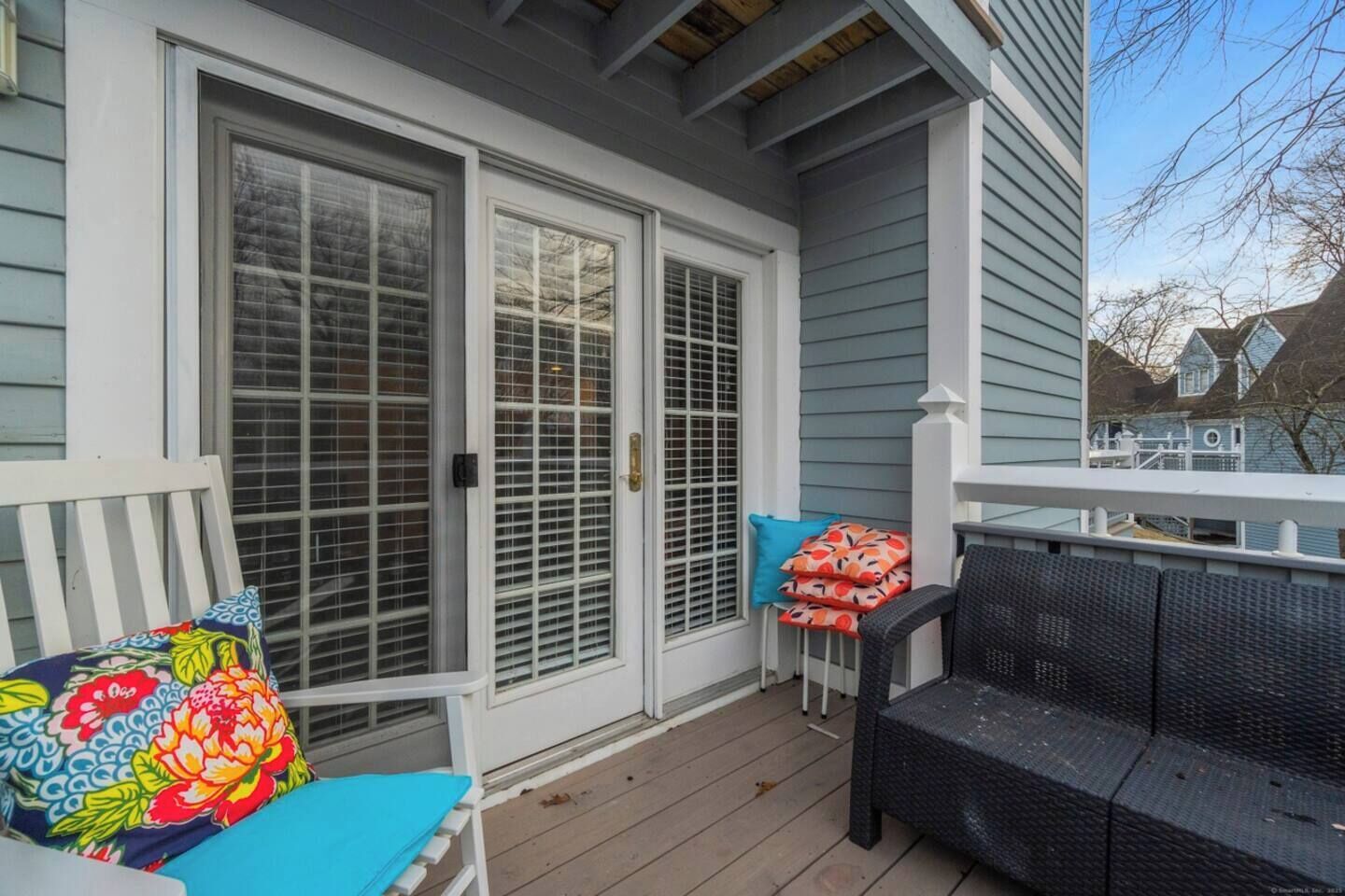 A covered porch with a white door, a bench with colorful cushions, and a small table with red and orange cushions.