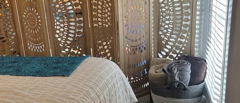 A hotel room with a bed, a blue throw blanket, a wicker basket with towels, and a decorative wooden partition with cut-out patterns.