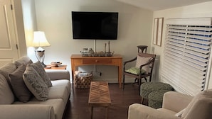 Een woonkamer met een bank, fauteuil en voetenbankje, een flatscreen televisie aan de wand en een houten salontafel.