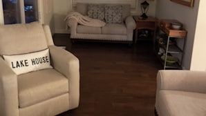 Ein gemütliches Wohnzimmer mit einem beigen Sessel, einer Couch und einem Holztisch. Eine Lampe und ein Spiegel stehen auf einem Beistelltisch.