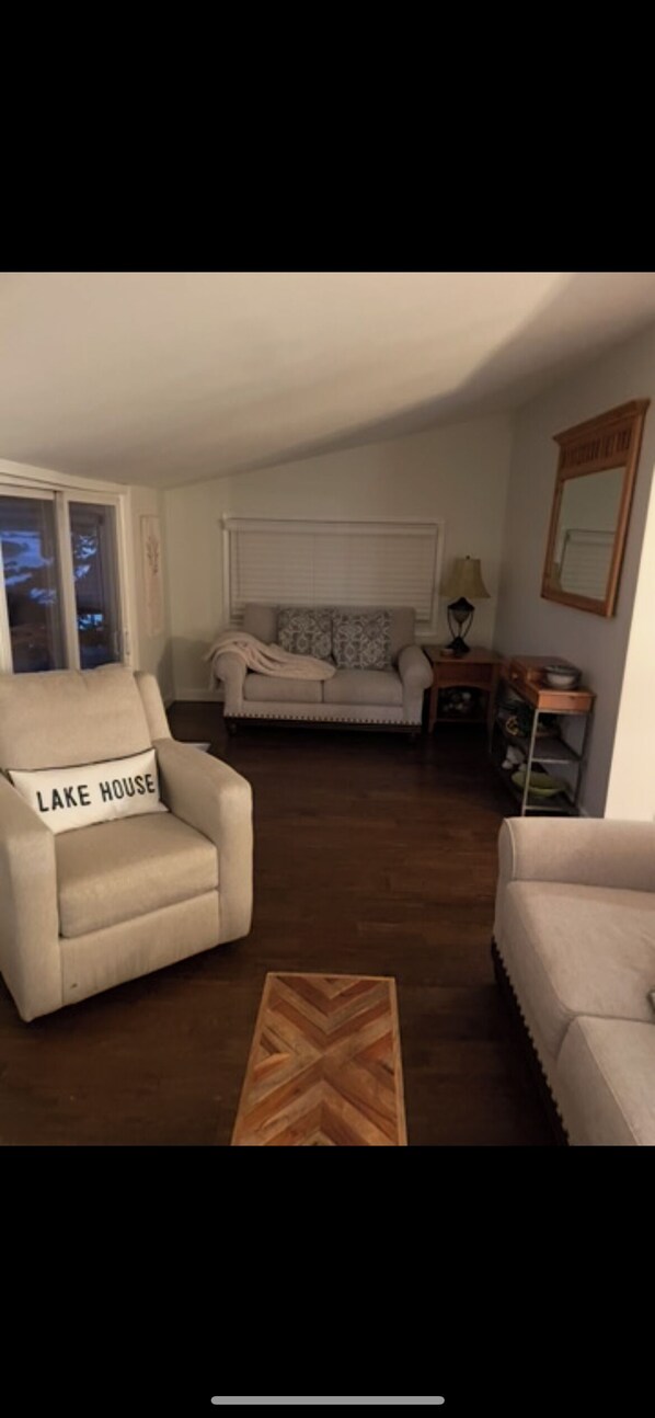 A cozy living room with a beige armchair, a sofa, and a wooden coffee table. A lamp and a mirror are on a side table.