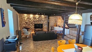 A rustic interior with a stone wall, wooden ceiling, a TV, a sofa, and a dining area with a table and chairs.