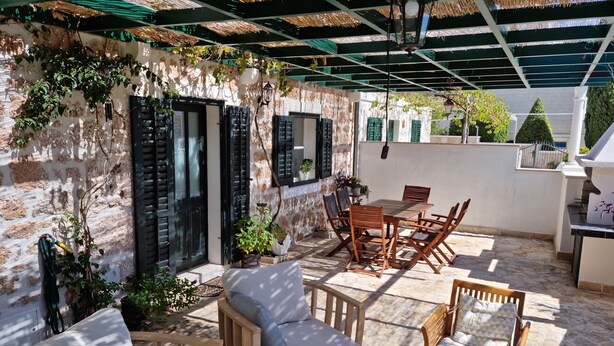 A covered outdoor seating area with wooden furniture, a table, and chairs, surrounded by potted plants and a stone wall.