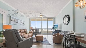 A spacious living room with a gray sofa, a brown ottoman, and a dining area with a wooden table and chairs. A ceiling fan and a large window with a view are visible. - Ocean Club 1106 | Enjoy Views of the Gulf! (Biloxi)