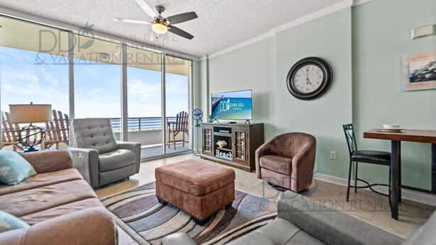 A spacious living room with a large window, a ceiling fan, a television, and a dining area. - Ocean Club 1106 | Enjoy Views of the Gulf! (Biloxi)