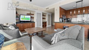 A modern living room with a grey sofa, a wooden coffee table, and a TV. The kitchen has wooden cabinets and a microwave. - Ocean Club 204 Deluxe | Stunning Panoramic Views of the Gulf! (Biloxi)