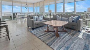 A spacious living room with a large window, a ceiling fan, and a dining area with a table and chairs. - Ocean Club 204 Deluxe | Stunning Panoramic Views of the Gulf! (Biloxi)