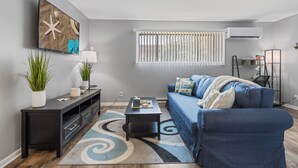 A living room with a blue sofa, a coffee table, a TV, and a large window with blinds. - Spacious2BR/Work Crews & Families/Sleeps6/desk/Parking/WIFI/Pet friendly (Aurora)