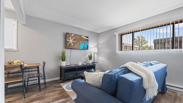 A living room with a sofa, a dining table with chairs, a TV mounted on the wall, and a window with blinds. - Spacious2BR/Work Crews & Families/Sleeps6/desk/Parking/WIFI/Pet friendly (Aurora)