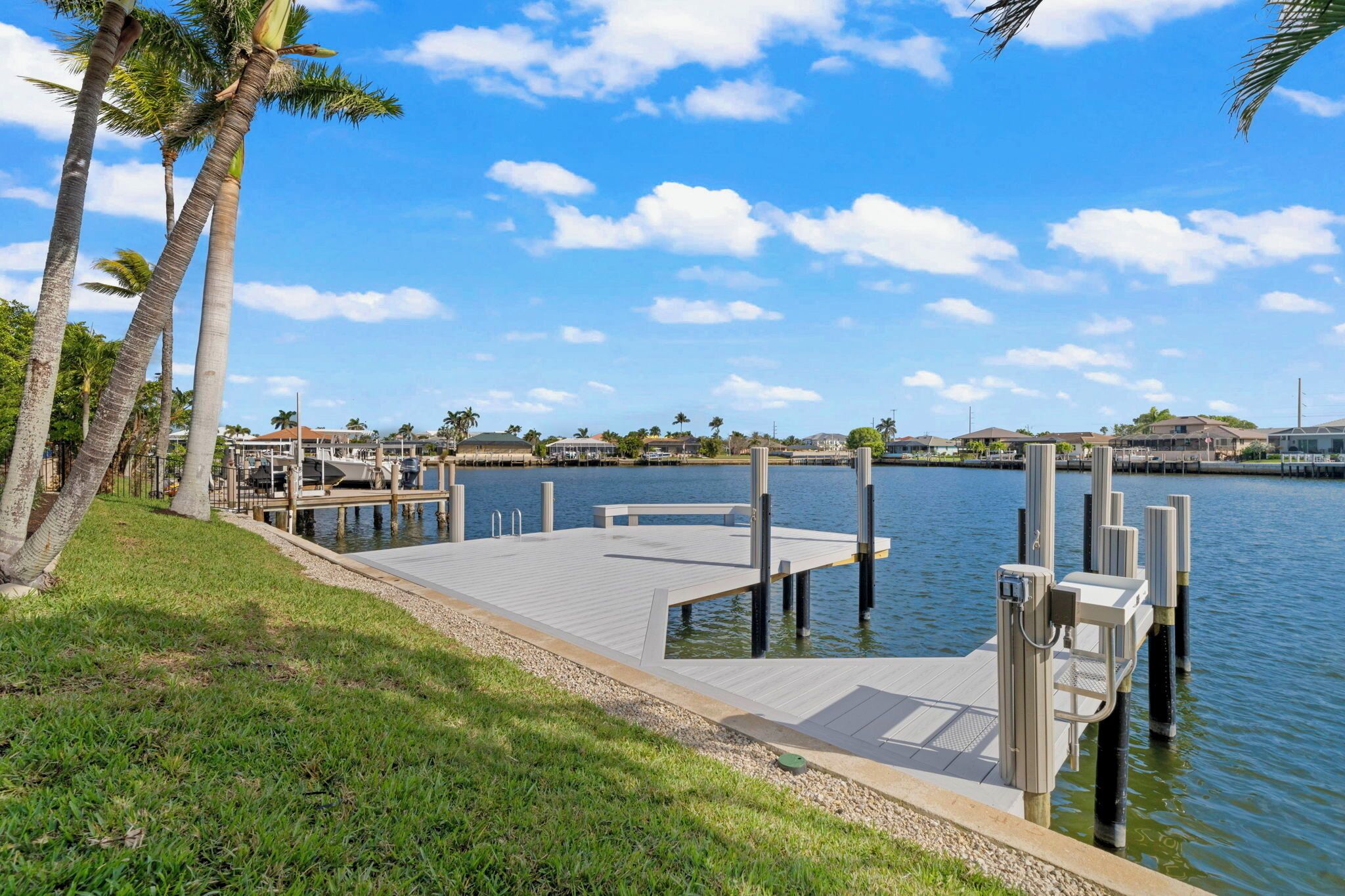 A waterfront dock with a grassy area, palm trees, and a clear blue sky.