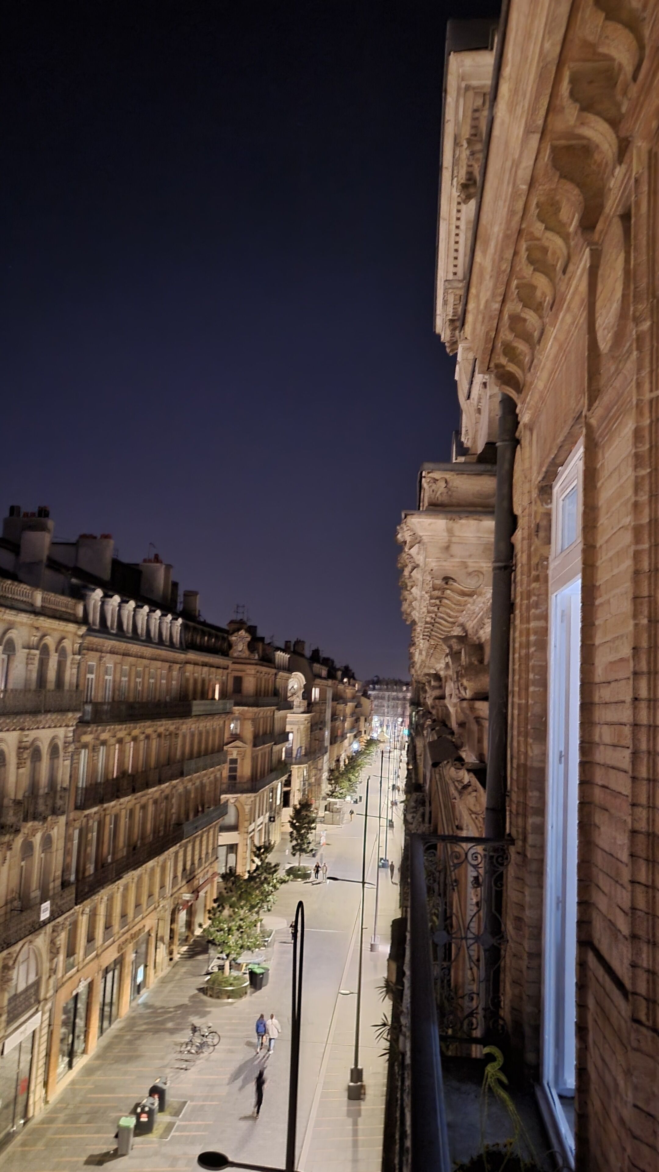 Vista nocturna de una calle con edificios históricos, una senda peatonal y algunas personas caminando.