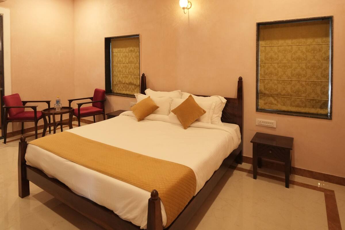 A hotel room with a large bed, a ceiling fan, a small table with chairs, and a window with a patterned curtain.