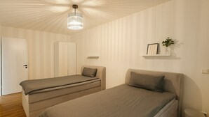 Une chambre avec deux lits simples, un luminaire au plafond, une petite plante posée sur une étagère et une porte.