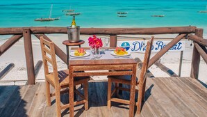 Una terraza de madera con una mesa dispuesta para dos, con vistas a una playa de aguas turquesas y barcos.