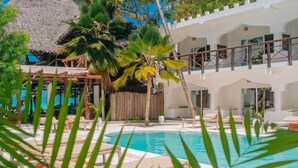Un área de piscina con sillones de descanso, una estructura de techo de paja y un edificio de dos pisos con balcones.