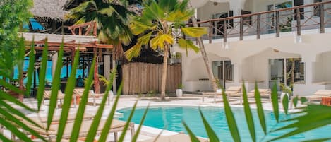 Un área de piscina con sillones de descanso, una estructura de techo de paja y un edificio de dos pisos con balcones.