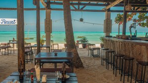 Un restaurante frente a la playa con mesas, sillas y un bar, con vista al océano.