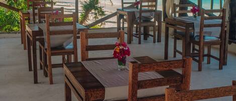 Un restaurant situé en bord de mer, avec des tables et des chaises en bois, un toit de chaume et une vue sur l’océan.