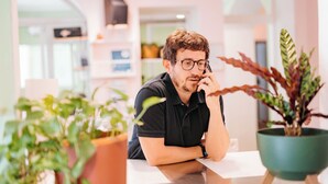 A person sitting at a table in a well-lit indoor space with potted plants and shelves in the background. - Tomato Backpackers (Turin)