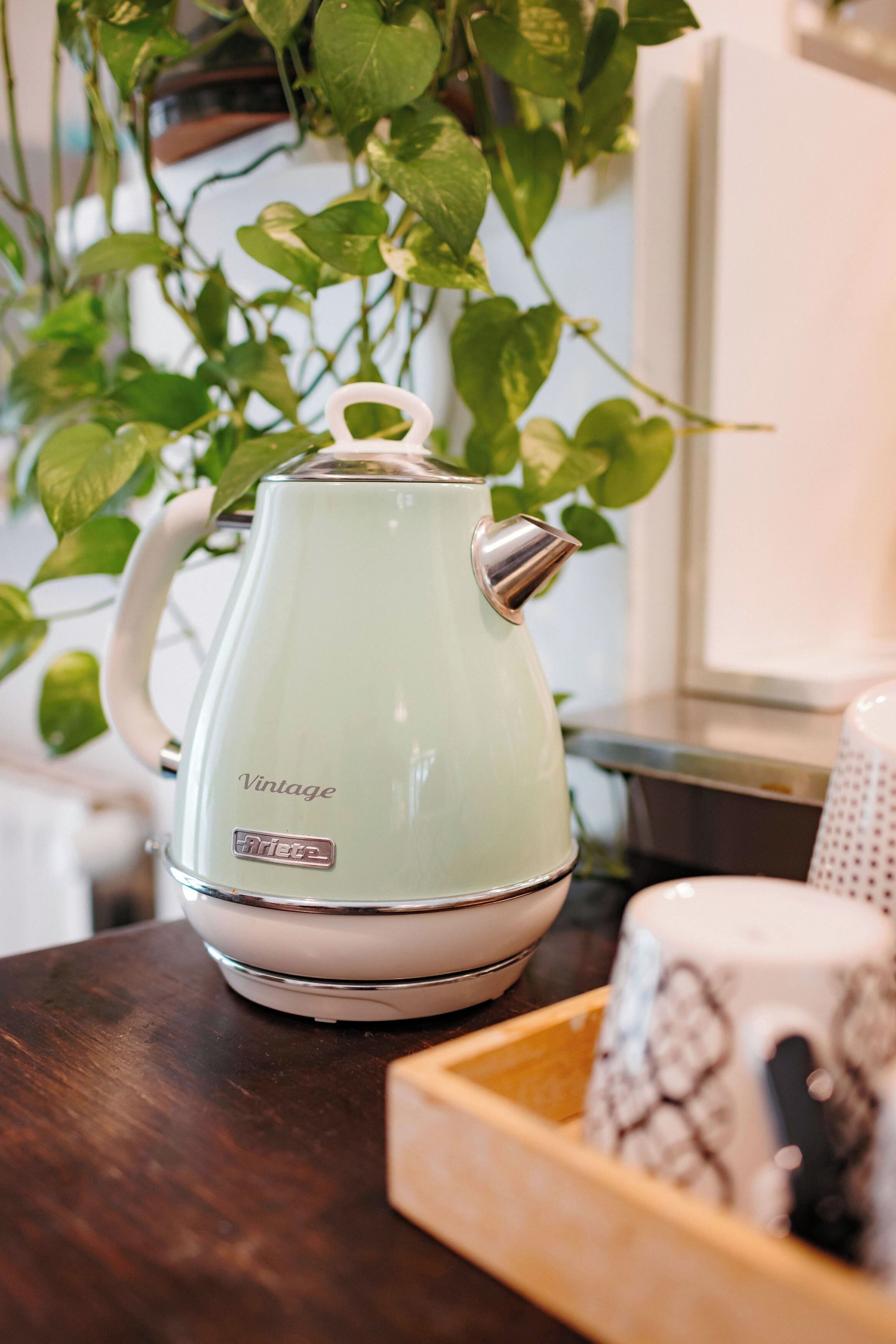 Una tetera de estilo vintage sobre una mesa de madera con una bandeja de objetos, incluyendo una taza estampada, en una habitación con una planta al fondo.