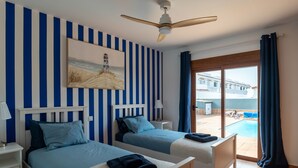 A hotel room with two beds, a ceiling fan, a painting of a lighthouse, and a view of a pool and building outside. - Villa Skawi - Cliffside Retreat (Costa de Antigua)