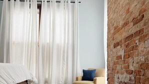 Une chambre à coucher avec un lit en bois, une chaise, un mur en briques et une fenêtre ornée de rideaux blancs.