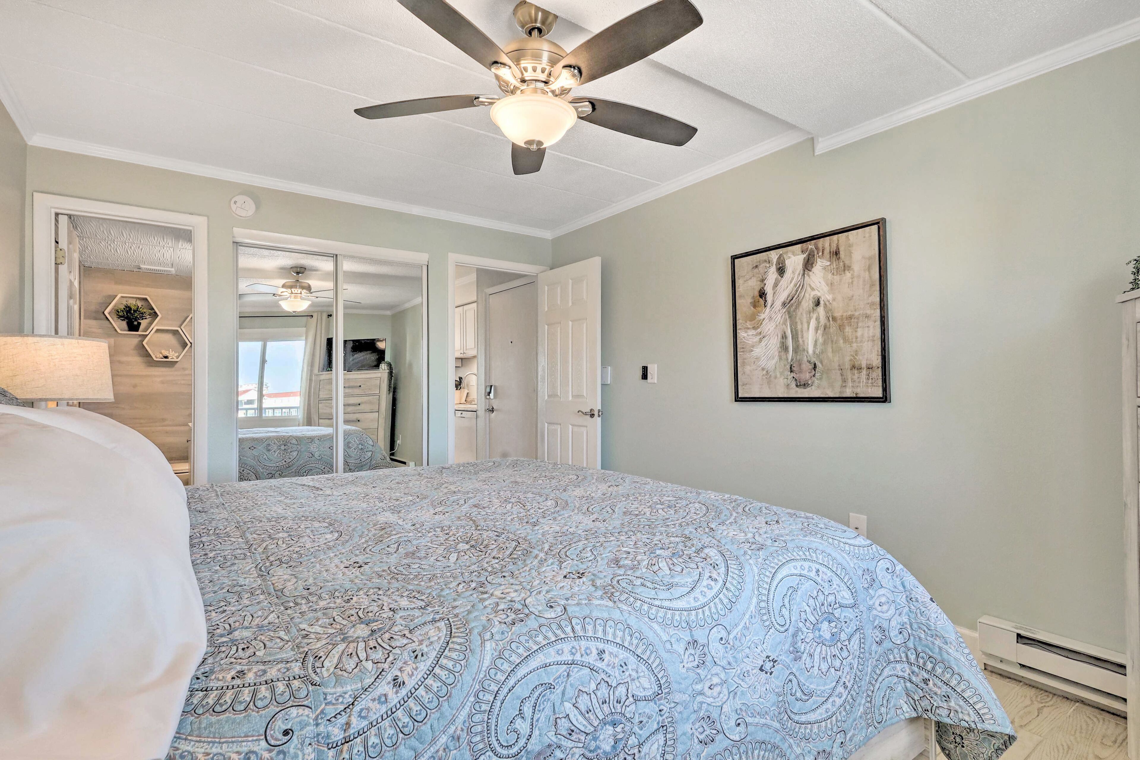 A bedroom with a ceiling fan, a bed with a patterned bedspread, a painting on the wall, and a hallway leading to another room.