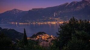 A coastal town at dusk with a mountain backdrop and a calm lake reflecting city lights.