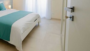 A hotel room with a bed, a nightstand, a lamp, a window with curtains, and an air conditioning unit. - Modern and functional space in the heart of the city (Mazara del Vallo)