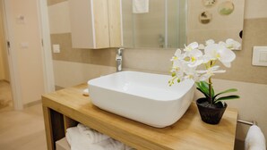 A bathroom with a white sink, a wooden vanity, a mirror, and a potted orchid. - Modern and functional space in the heart of the city (Mazara del Vallo)