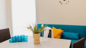 A dining area with a table set for four, a vase with flowers, and a blue sofa with yellow pillows in the background. - Modern and functional space in the heart of the city (Mazara del Vallo)